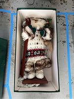 Alaskan Santa figurine dressed in white Nordic patterned sweater with fur trim, holding red skis while standing on a wooden base inside original box.