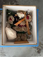 Top-down view of Woodsman Santa figure wearing fur-trimmed clothes holding wooden skis, ski poles, a small Christmas tree, and bear figure in original box.