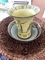 Yellow Art Nouveau style vase with black classical figures inside nested patina metal bowls and large wicker basket beneath.