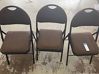Three brown padded Cosco folding chairs set up side by side on a concrete floor, showing front and side views