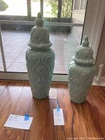 Two teal decorative ceramic ginger jars side by side on wooden floor next to glass door. Both have lids with finials and embossed patterns.