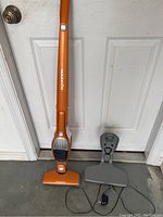 Orange Electrolux Ergorapido cordless stick vacuum with charging station and power adapter set in front of a white door.