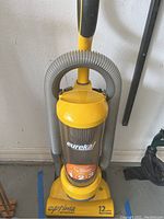 Full frontal view of yellow Eureka Optima upright vacuum with hose wrapped around the handle and clear canister showing the brand and model details.