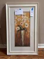 Photo of floral art print featuring white orchids in a tall vase, cream-colored frame, and white matting on a beige wall.