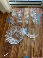 Ten clear glass mugs arranged in two rows with white snowflake designs visible. Two glass bowls placed below the mugs: one large cut glass bowl with swirl pattern, and a smaller glass bowl beside it with scalloped edges.