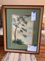 Framed watercolor painting showing a solitary pine tree with grass and cloudy sky. The frame is wooden with a green mat border. A tag with measurements and lot number is visible.