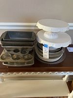 Photo showing multiple metal loaf pans and muffin tin stacked beside two white ceramic cake stands and a stack of glass plates.