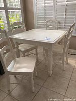 Wide view of white square kitchen table with four matching chairs around it in kitchen area, showing overall set and condition.