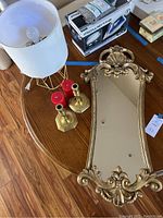 Top view of ornate gold-tone mirror with curved shape, visible crack in frame, beside modern lamp with geometric gold-tone base and white shade, pair of brass candlesticks with red candles on wooden table