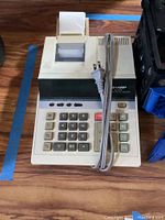 Sharp printing calculator on wood surface, showing keypad and receipt tape.
