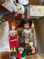 Image showing Little Orphan Annie doll, multiple other vintage dolls inside a box along with stuffed bunny, playing cards, and ceramic fish visible nearby.
