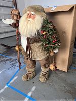 Full view of the vintage Santa figure in woodsman attire holding a decorated Christmas tree and a rustic wooden staff with birdhouse, set on a garage floor with original box behind.