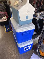 Three coolers stacked: one Igloo Playmate on top in blue and white, two blue Coleman coolers underneath with white lids and handles.