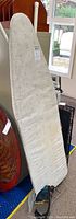 Padded ironing board with white cover and metal legs standing upright indoors near a window.