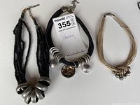 Photo showing three beaded necklaces including black multi-strand with large silver metal rings, black multi-strand with silver discs, and natural fiber necklace with a knot and small silver bells.