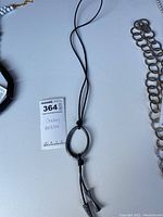 Photo showing the full black cord necklace with a silver-tone textured oval pendant and two dangling bars, laid out flat on a white surface.