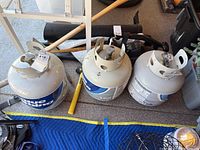 Three Blue Rhino propane tanks placed side by side on the ground with various items around them.