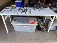 White rectangular collapsible table with various items displayed on top and large storage bin underneath.