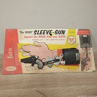 Front view of The Secret Sleeve-Gun original cardboard packaging with black plastic gun, metal articulating arm, and bullet storage.