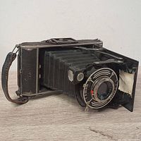 Front view of vintage folding camera showing extended black bellows, circular shutter dial, lens, and leather strap.