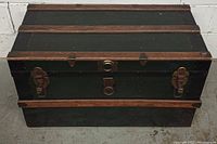 View of closed antique trunk showing exterior with black finish, wooden slats, and metal clasps on front.