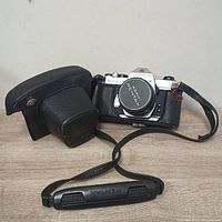 Front view of Asahi Pentax Spotmatic F camera with lens cap on, leather carrying case beside it and black camera strap with Pentax branding laid out on tabletop.