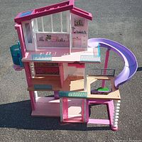 Front and side view of the Barbie Dream House showing three floors, open walls with printed interior pictures, blue balcony railings, and purple slide