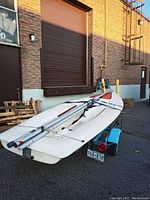 Aft view: white fiberglass sailboat hull loaded on light blue single-axle trailer with folded aluminum mast and boom strapped on deck