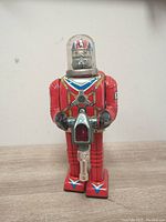 Front view of red Cragstan X-70 Astronaut tin-litho robot with clear plastic dome and transparent ray gun, showing printed blue, white, and silver accents.