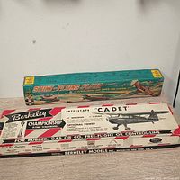 Two model kit boxes stacked; visible text shows Berkeley Interstate 'Cadet' flying scale model and colorful stunt-flying plane box.