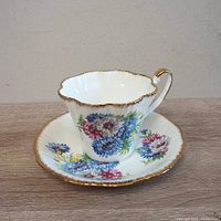 Front side view showing the floral pattern on white bone china teacup and matching saucer with scalloped gold-trimmed edges