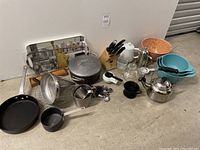 Photo showing assortment of kitchen items including cookware, mixing bowls, measuring spoons, teapots, serving tray, knife block and utensils.