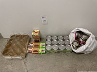Sets of clear glass jelly jars in a cardboard tray, boxes of new jar lids, and a white bag full of metal lid rings on the floor against a wall.