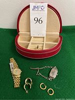 All items displayed on green fabric: red travel case, earrings, ring, and two watches.