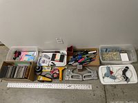 Whole assortment of office supplies and household items arranged on the floor including pens, clipboards, staplers, nails, a hot glue gun, and locks.