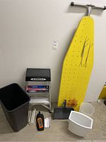 Photo showing the yellow ironing board with clips and rust, large dark trash can, two small trash cans, brand new dustpan, step ladder, surge protector, and digital sound level meter.