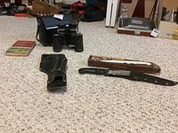 Full view of the lot including binoculars with carrying case, leather holster, hunting knife in sheath with original box, and fishing book.