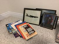 Overall lot showing the stack of travel and aviation books on the floor and four framed jet-related prints leaning against the wall.