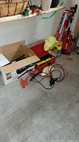Garage floor showing red and black booster cables with clamps, small red snow shovel, and the edge of a red toolbox.