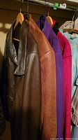 Four coats hanging on hangers showing brown leather coat, brown/tan jacket, bright purple and deep pink zip-up jackets.