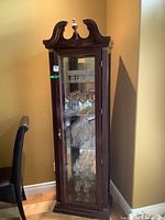 Full view of the tall curio cabinet showing dark wood finish, glass door, and displayed glassware inside.