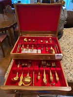 Open wooden canteen showing two drawers filled with gold-plated cutlery pieces arranged neatly within red felt lining. Visible various forks, spoons, and serving utensils.