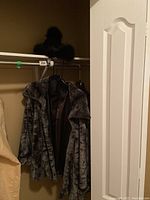 Full view of hanging gray faux fur jacket with wide collar and black interior lining, along with knit hat on the rack.