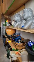 Shelving with two concrete angel head garden decorations, flower pots, colorful glass garden balls, metal basket with garden tools, hose brush attachment and watering can