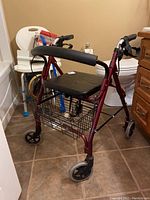 Maroon folding rollator with black padded seat, wire basket underneath, and ergonomic handles with brakes, photographed in bathroom with other medical items around