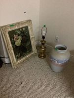 Floral print leaning against wall beside the metal table lamp and the pottery vase on carpeted floor