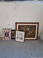 Four framed artworks on floor against wall, showing variety of styles including floral needlepoint, animal prints, and canvas painting.