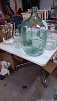 Five glass jugs of various sizes on a white table, including green tinted and clear glass jugs.