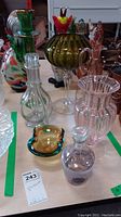 Photo showing full set of glassware including a green and gold tall vase, pedestal bowl, pink ribbed vase, amber ashtray, and clear decanter on a table.