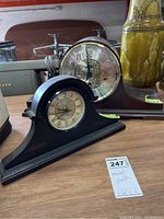 Two mantel clocks displayed on a wooden surface with a price tag, showing the smaller clock with Roman numerals in front and the larger clock behind.